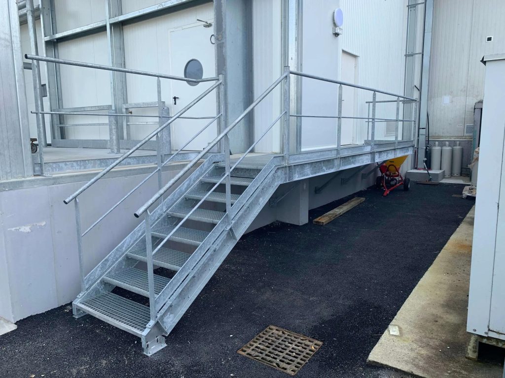 Horganyzott kültéri acéllépcső rácsos járófelülettel, korláttal és pihenőplatformmal ipari épület mellett.